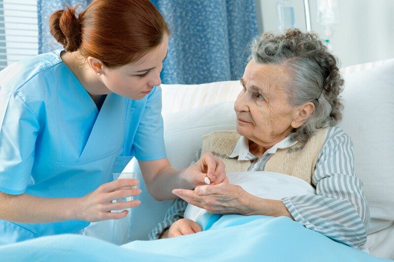Krankenpflegerin am Krankenbett einer älteren Dame, der sie Medikamente verabreicht