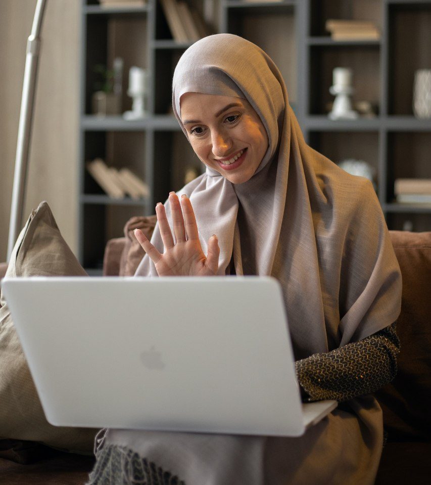 Frau mit Hijab sitzt auf Couch vor Bücherregal. Sie winkt. Videokonferenz. Sie hat einen Laptop auf dem Schoß