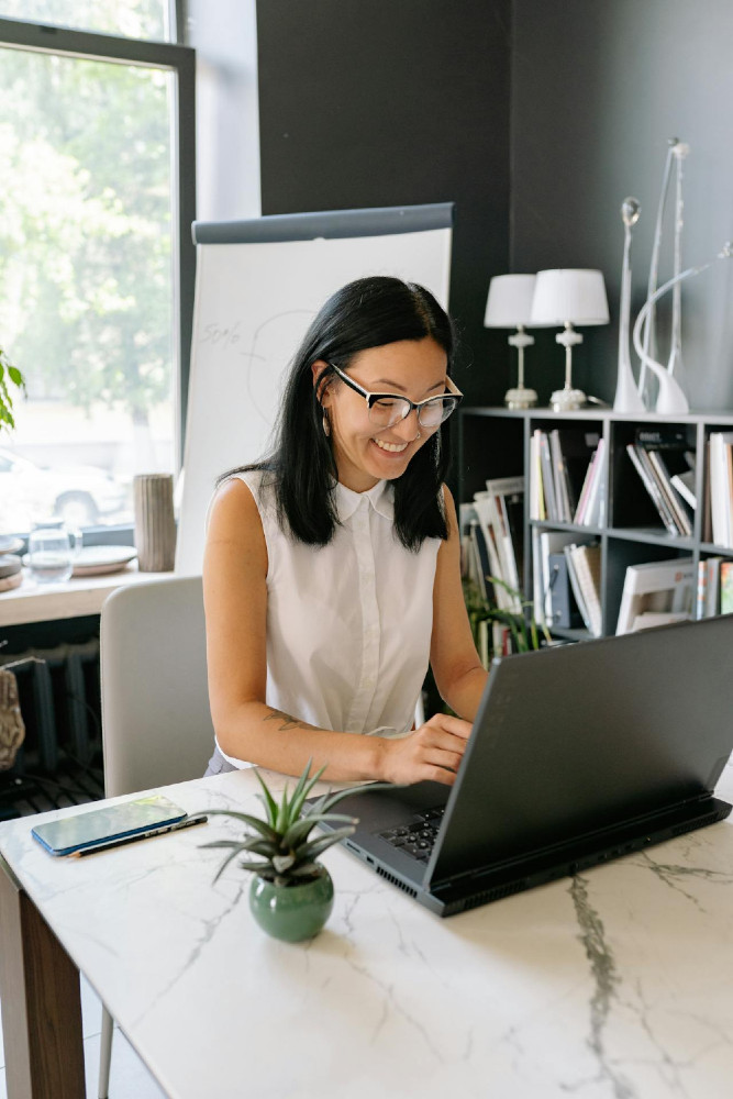 Asiatische, lächelnde Frau sitzt an einem Schreibtisch und arbeitet an einem Laptop. Hinter ihr ist eine Flipchart und ein Fenster, plus Regale voller Akten. Sie ist in einem Büro.